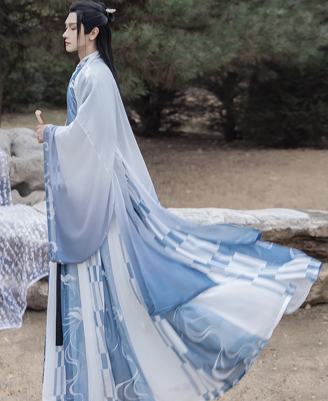 Side profile of male model in flowing blue and ivory Wei Jin hanfu for men featuring wide sleeves, abstract printed skirt, black waist ties, and folding fan