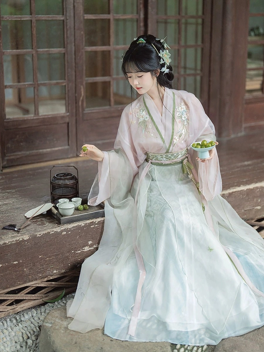 Three-quarter view of a Wei Jin Dynasty Hanfu outfit featuring floral embroidered pink upper robe, green collar trim, light cyan wrap skirt, and pink skirt edging beside a traditional tea setting
