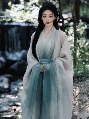 Front view of a woman wearing a layered Wei Jin Dynasty Hanfu robe in a creamy white and teal green gradient, featuring a cross-collar V-neck, sheer outer layer, wide sleeves, and matching waist sash near a forest waterfall