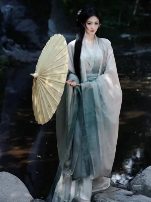 Full-body side view of a woman wearing a Blue Moon Lake Wei Jin Dynasty Hanfu robe with creamy white and teal gradient fabric, wide flowing sleeves, high-waisted skirt, waist sash, and a bamboo oil-paper umbrella beside a woodland stream