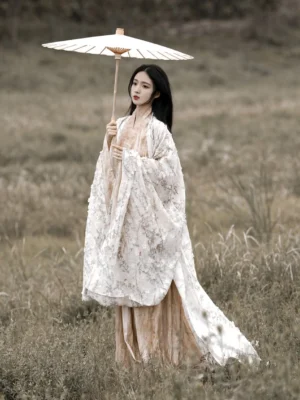 Front three-quarter view of a woman wearing a Wei Jin Dynasty Qiyao Ruqun Hanfu with a floral big sleeve coat beige inner robe flowing skirt and orange waist sash in a grassy field
