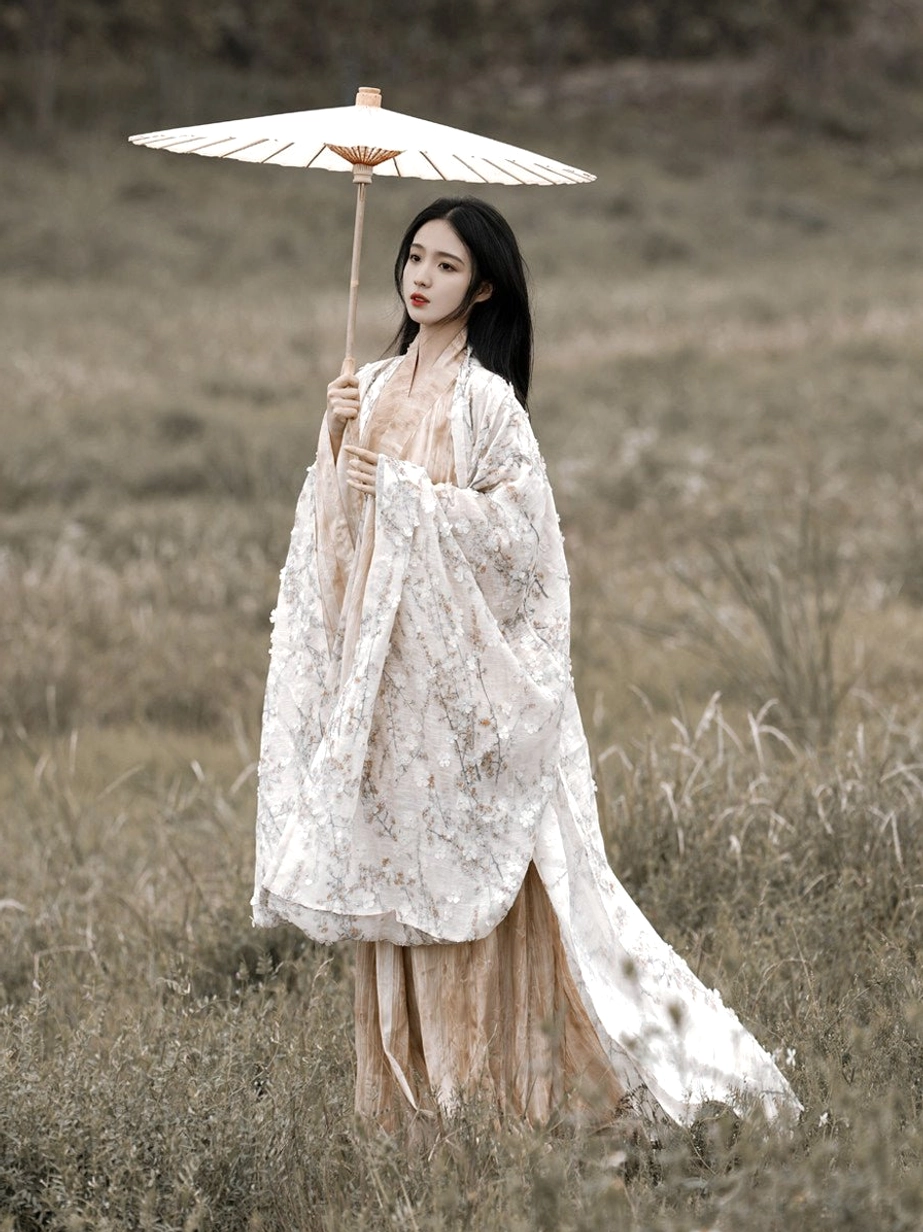 Front three-quarter view of a woman wearing a Wei Jin Dynasty Qiyao Ruqun Hanfu with a floral big sleeve coat beige inner robe flowing skirt and orange waist sash in a grassy field
