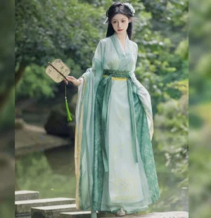 Full-body front view of a jade-green Wei Jin Dynasty Qiyao Ruqun Hanfu with wide flowing sleeves, mint and dark green layers, cross-collar top, and yellow waist trim beside a woodland waterside path