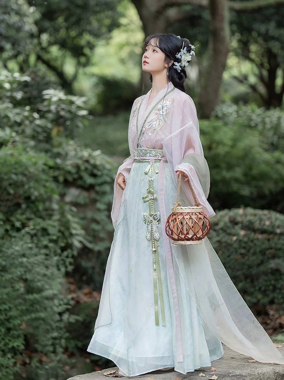 Wei Jin Dynasty Qiyao Ruqun Hanfu in blush pink and light cyan with pine green trim, floral embroidery, flowing sheer sleeves, and decorative waist ornaments photographed in a traditional Chinese garden