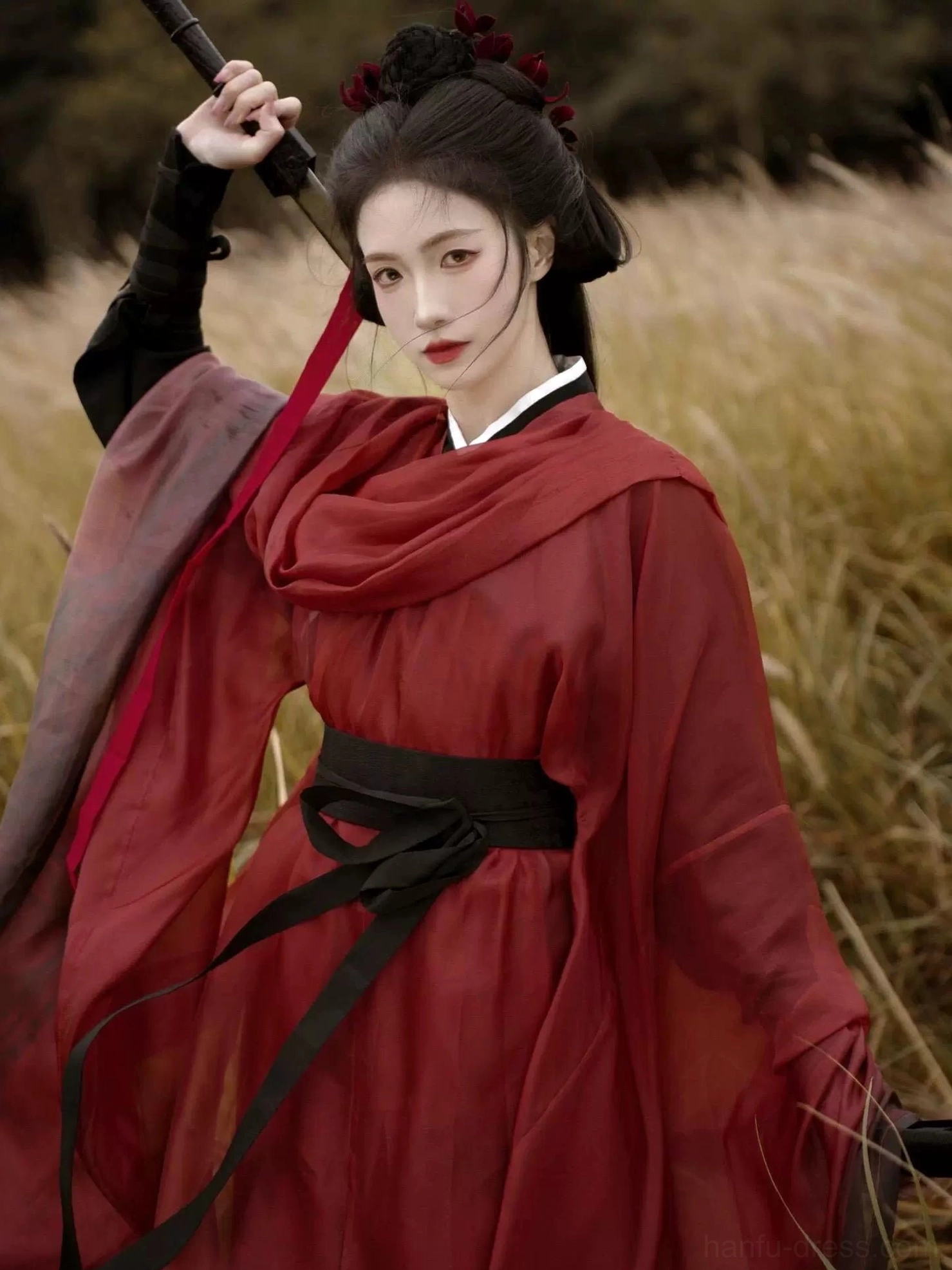 Front three-quarter view of a red and black Wei Jin Dynasty Qiyao Ruqun Hanfu with sheer dark red robe, black sash, cross collar, and flowing sleeves in an autumn reed field
