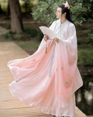 Male model wearing Wei Jin inspired pink ombre Qiyao Ruqun hanfu set with flowing wide sleeves and light blue tassel waist sash on a waterside wooden path