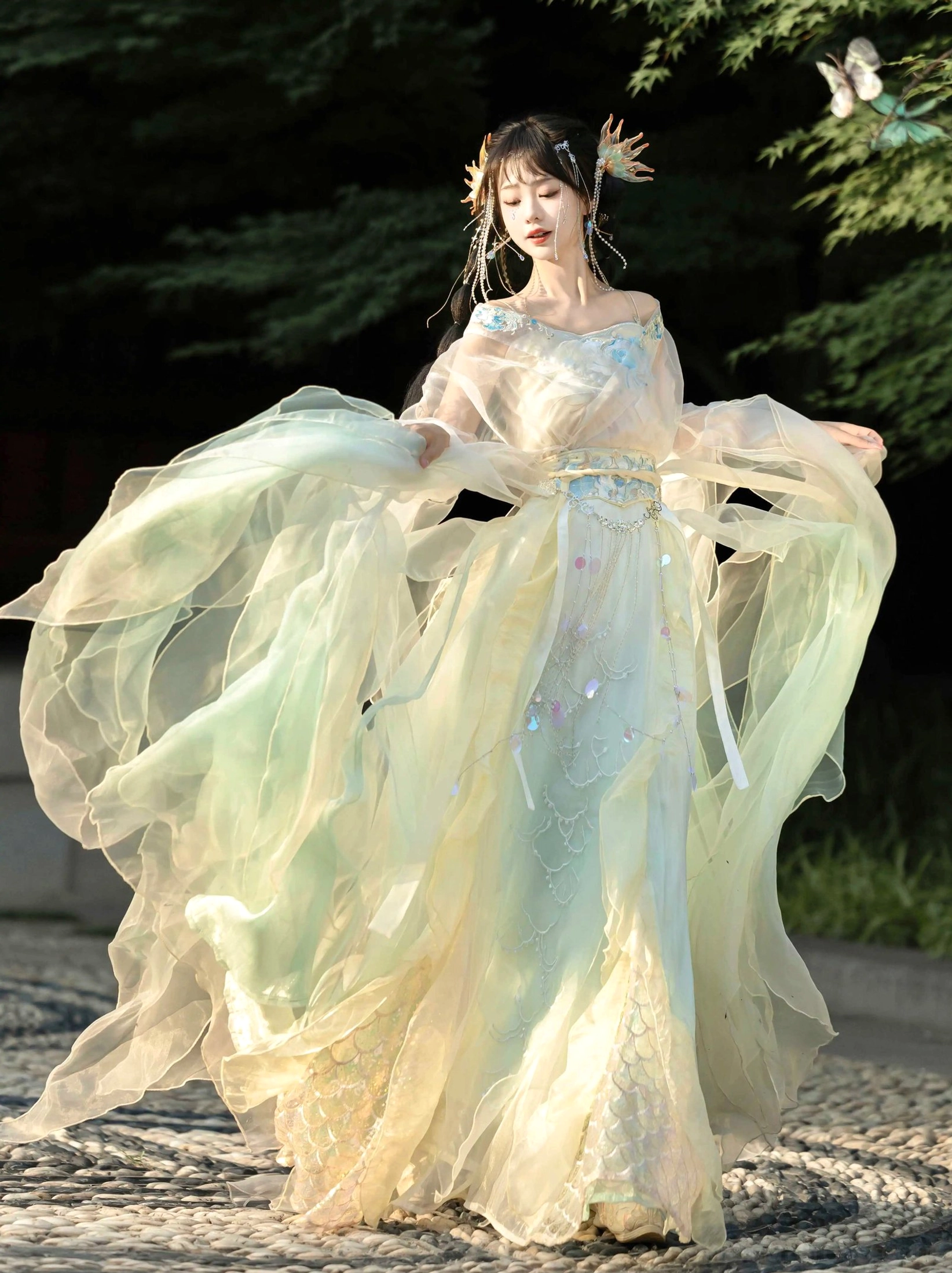 Light yellow green gradient Wei Jin Dynasty Qiyao Ruqun Hanfu with off shoulder top flowing sheer wide sleeves beaded waistband and fish scale skirt embroidery worn in a traditional Chinese garden