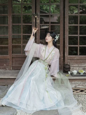 Front view of a Wei Jin style Qiyao Ruqun Hanfu with pink embroidered top, light cyan pleated skirt, green collar details, sheer sleeves, and silk round fan in a traditional courtyard