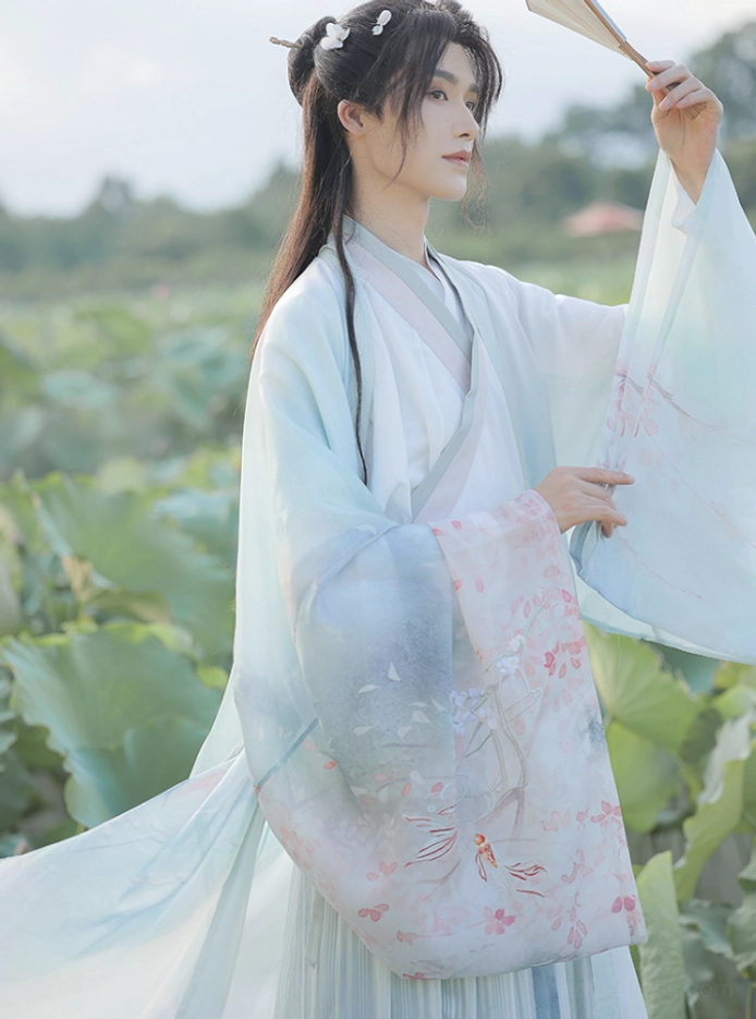 Side three-quarter view of a male model in traditional Chinese wuxia hanfu with a flowing long hem, embroidered cuffs, pastel layers, hair ornaments, and folding paper fan