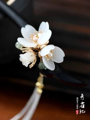 Close-up angled view of a traditional Chinese hair stick with white blossom flowers, gold-tone accents, green jade-style beads, and a long layered beaded tassel on a black wooden stick