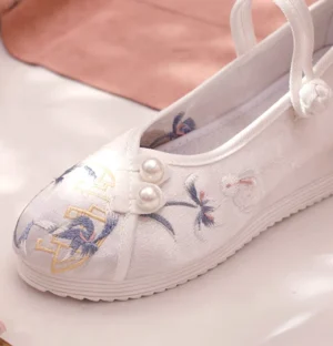 Close-up of a white satin Chinese traditional Hanfu shoe with blue flower and crane embroidery, golden motifs, pearl fasteners, Chinese knot ankle strap, and hidden height-increasing rubber wedge sole