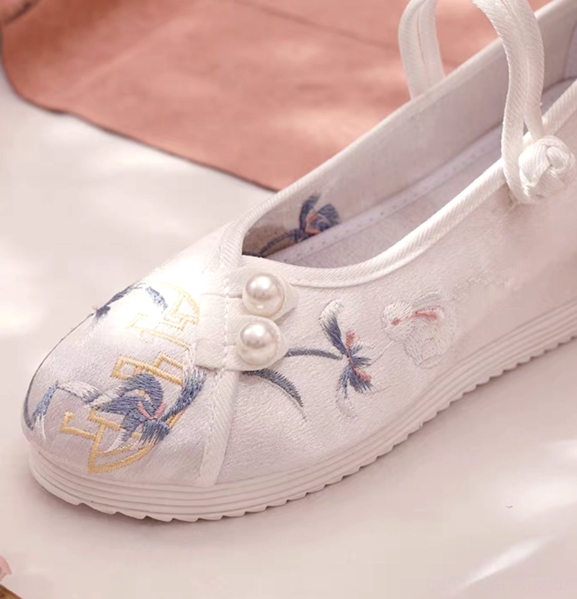 Close-up of a white satin Chinese traditional Hanfu shoe with blue flower and crane embroidery, golden motifs, pearl fasteners, Chinese knot ankle strap, and hidden height-increasing rubber wedge sole