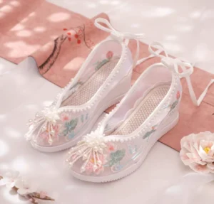 White embroidered Chinese Hanfu shoes with pink lotus flowers green leaves pearl tassels ankle tie straps and wedge platform soles displayed with pink fabric and flowers