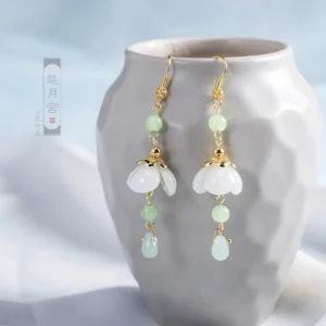 White glass flower Chinese dangle earrings with green beads and pale green teardrop pendants displayed on an embossed gray ceramic vase for Hanfu styling