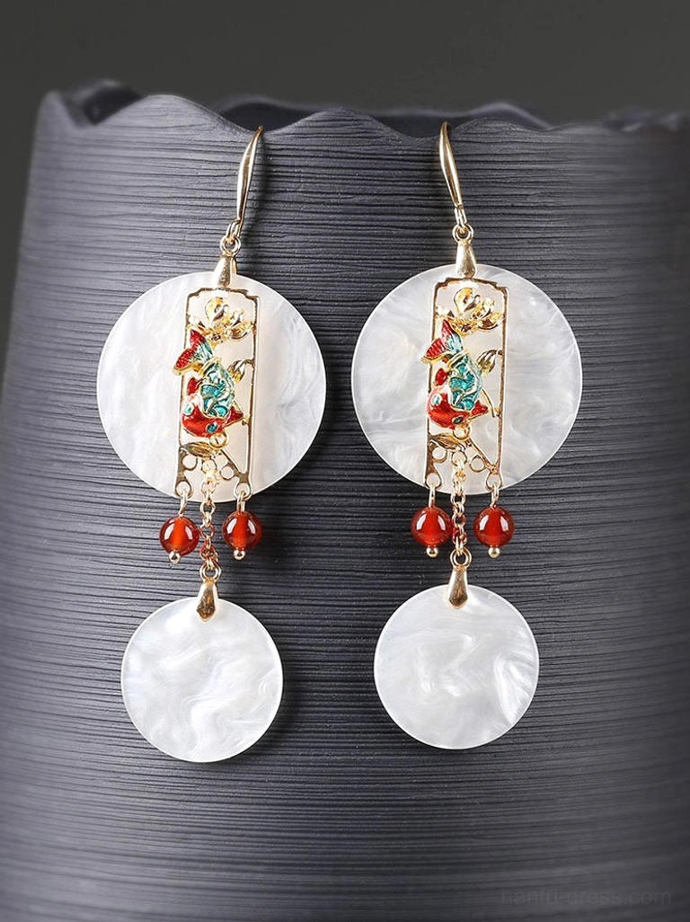 Close-up of white shell dangle earrings on a dark textured ceramic stand showing natural shell grain and red bead accents