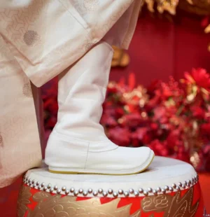 Side profile of a white traditional Chinese Hanfu boot with an upturned toe and tan sole edge displayed on a decorated Chinese drum stool beside festive red flowers