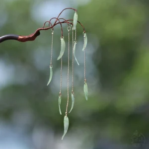 Traditional Chinese willow leaf hair stick with dark brown wooden handle, copper branch details, jade-green leaf pendants, and golden chain tassels against blurred green foliage