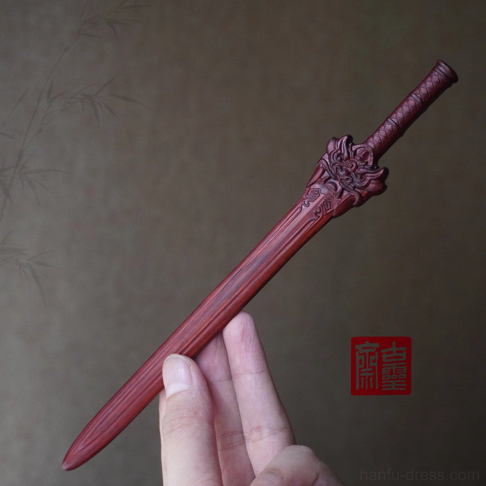 Wine red Chinese sword hair stick held in a hand to show its scale, featuring smooth carved wood texture, a mythical beast head, and detailed blade engraving