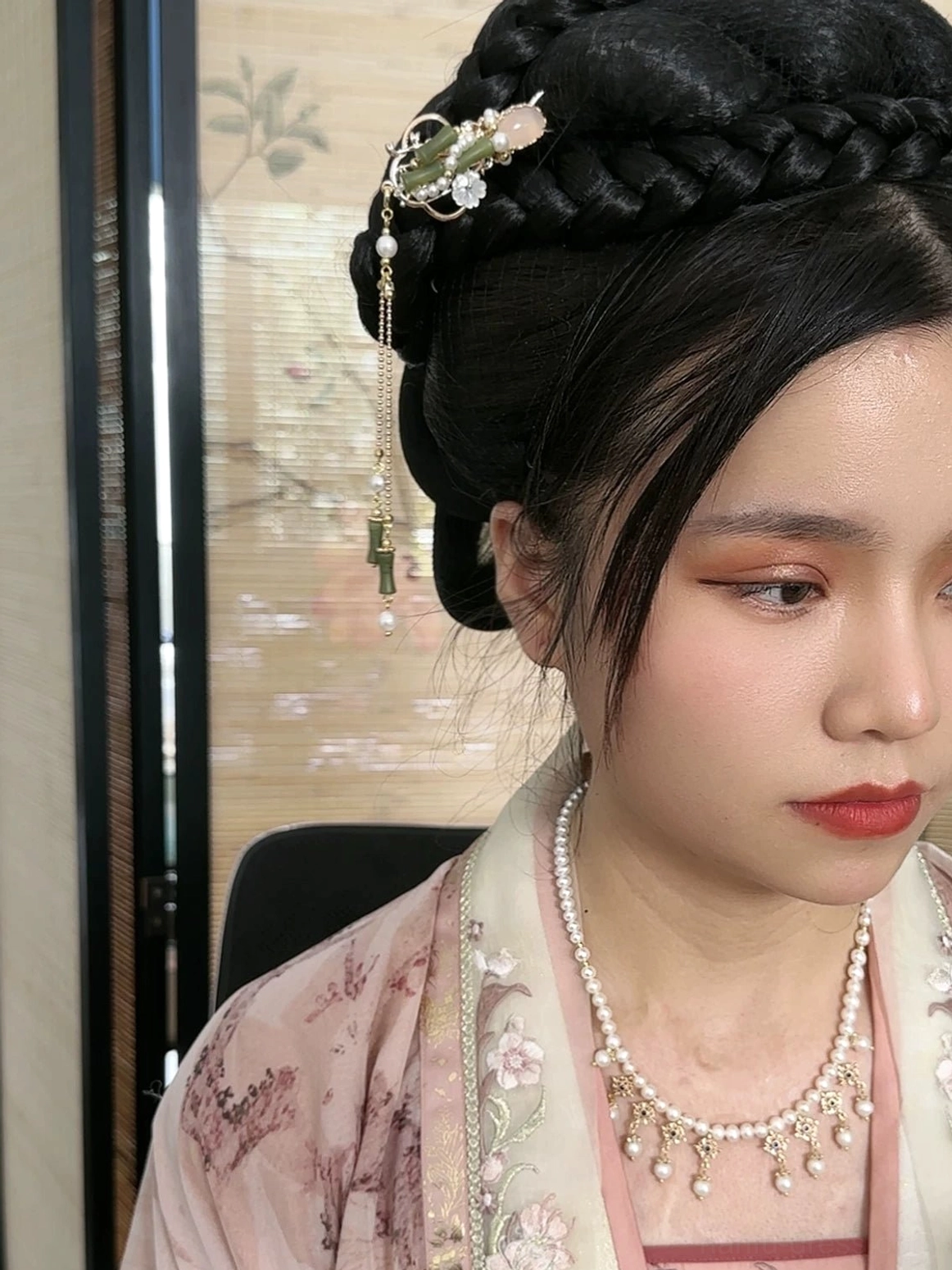 Woman wearing a bamboo tassel Chinese hair stick in an elaborate traditional hairstyle with embroidered Hanfu and layered pearl necklaces against a Chinese ink painting screen