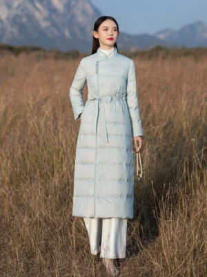 Full front view of a women's dusty blue Chinese style over knee down coat with stand collar frog buttons adjustable waist sash and horizontal quilting worn with white wide leg pants outdoors