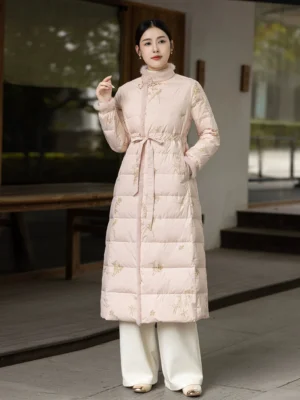 Pale pink women's Hanfu-style long down coat with fuzzy stand collar, golden bamboo and bird embroidery, quilted silhouette, and adjustable waist shown in a traditional Chinese courtyard
