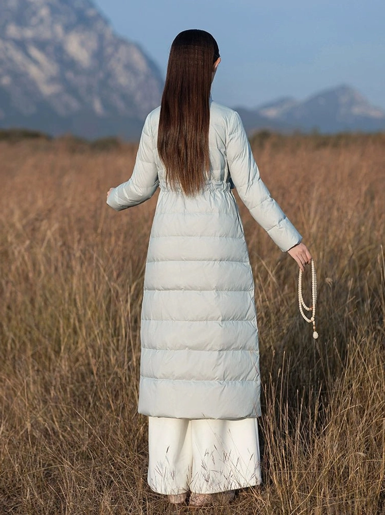 Back view of a women's long Chinese style quilted down coat in light dusty blue with stand collar waist sash and over knee winter silhouette
