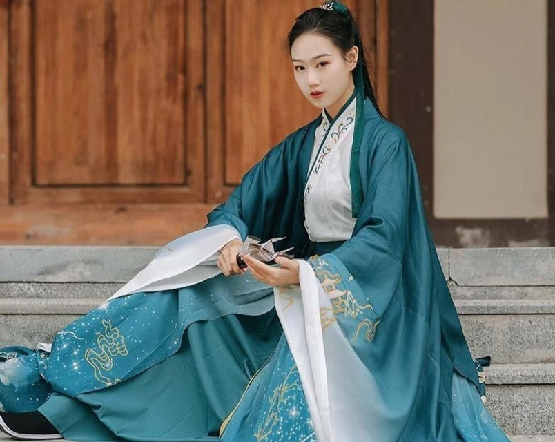 Wuxia style hanfu ruqun showing the neat cross-collar top, subtle blue gradient skirt, and embroidered details while seated beside an ancient wooden door