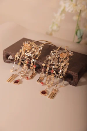 Pair of Xianglian traditional Chinese Hanfu hair pins with gold-toned openwork floral carving, burgundy gemstone accents, layered tassel pendants, dark wooden display stand, and white flower decoration