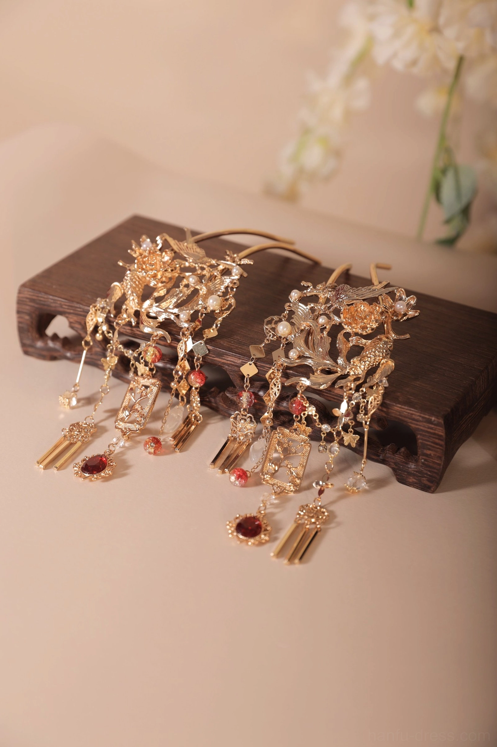 Pair of Xianglian traditional Chinese Hanfu hair pins with gold-toned openwork floral carving, burgundy gemstone accents, layered tassel pendants, dark wooden display stand, and white flower decoration