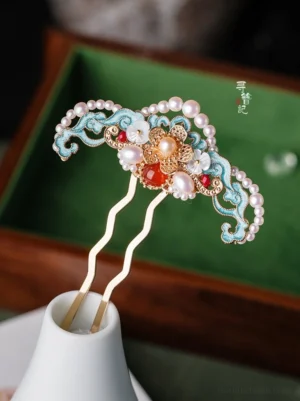 Close-up of a blue cloisonne Chinese Hanfu hair pin with cloud patterns, freshwater pearls, red agate, shell flower ornaments, and a gold-toned double-pronged stem displayed in a decorative holder