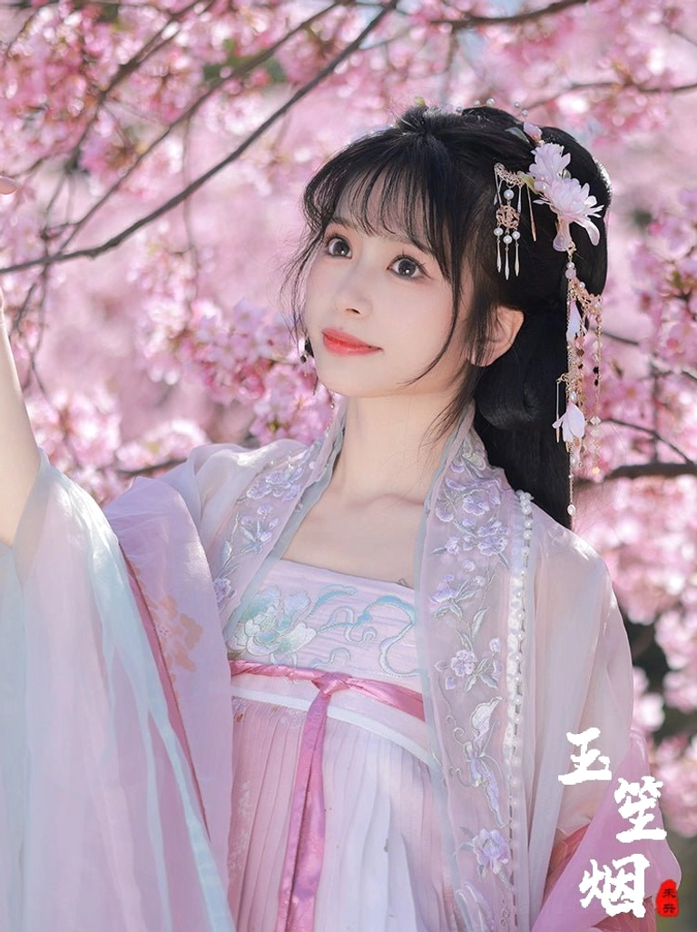 Front three-quarter portrait of a model wearing the Yuyue pink floral Hanfu hair accessory set with white flower ornaments and delicate tassels in a cherry blossom garden