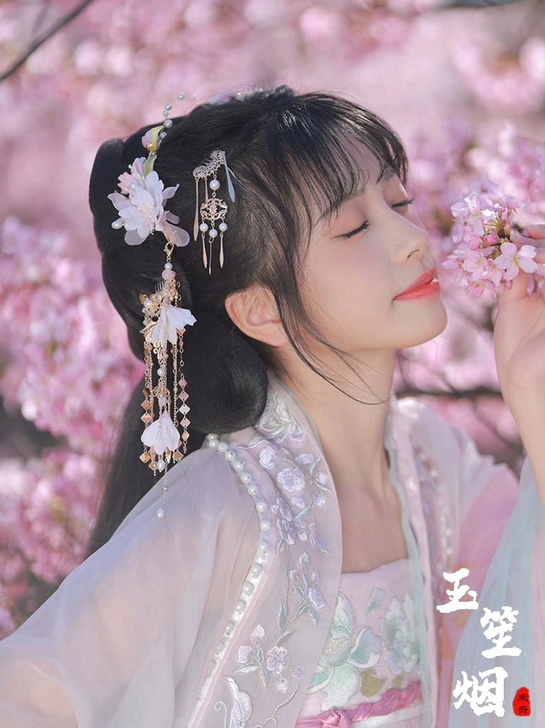Side profile model wearing soft pink Hanfu with Yuyue white flower hair clips and long tassel hair ornaments against cherry blossoms