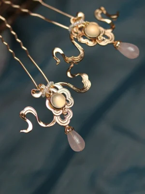 Zheyue traditional Chinese Hanfu hair pin with polished gold cloud filigree, light central stone, pink teardrop pendant, and slender wavy hair stick on dark blue fabric