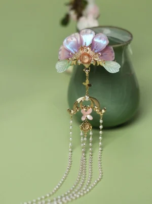 Zixuan traditional Chinese Hanfu hair pin with iridescent purple pink glass flower petals green resin leaves gold tone metal frame and long pearl tassel displayed beside a celadon vase